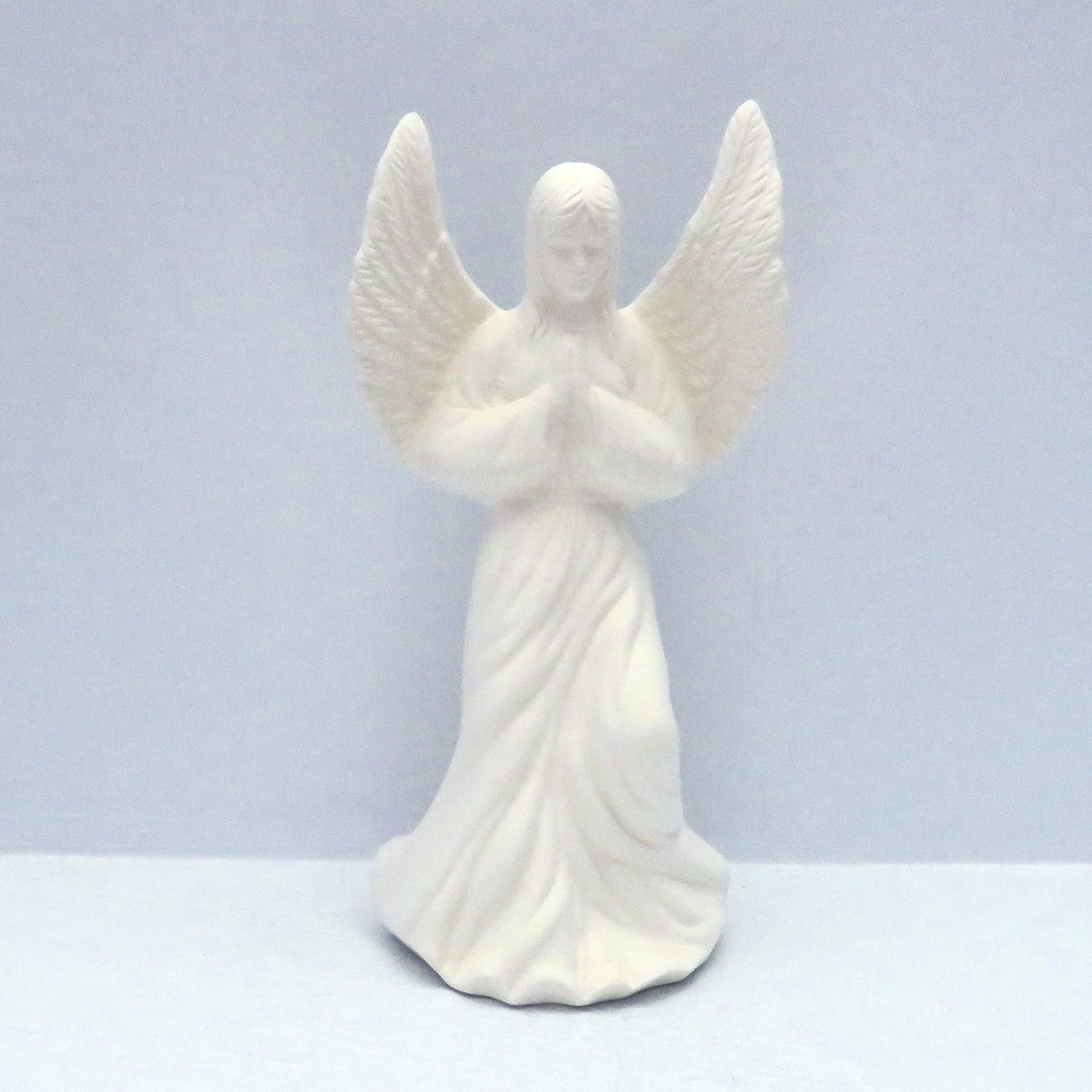 Unpainted ceramic praying angel figurine on a pale blue surface.  Her wings are outstretched and her head is slightly bowed.