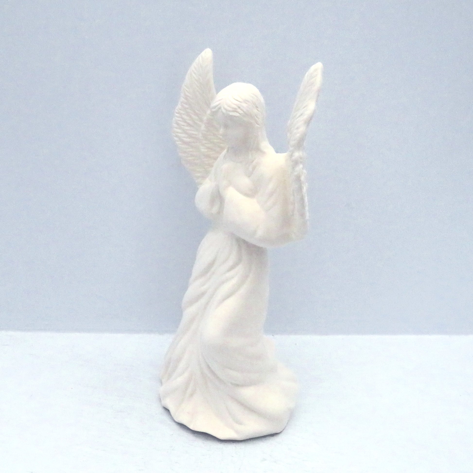 Side view of paintable ceramic angel figuine with wings outstretched and her hands in the prayer position.  She is against a pale blue background.
