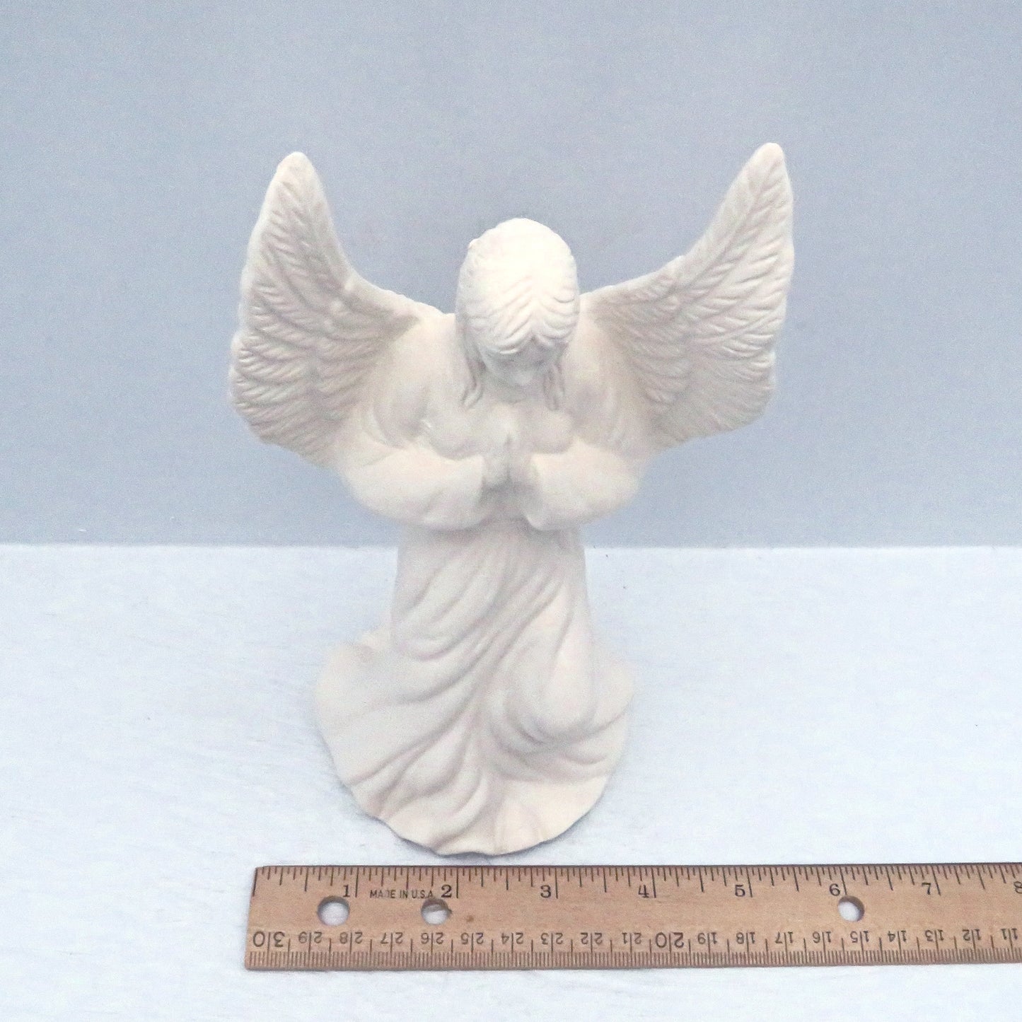 Top view of handmade ceramic praying angel figure near a ruler showing that she is approximately 3 3/4 inches across the wing span.  She is standing on a light blue background
