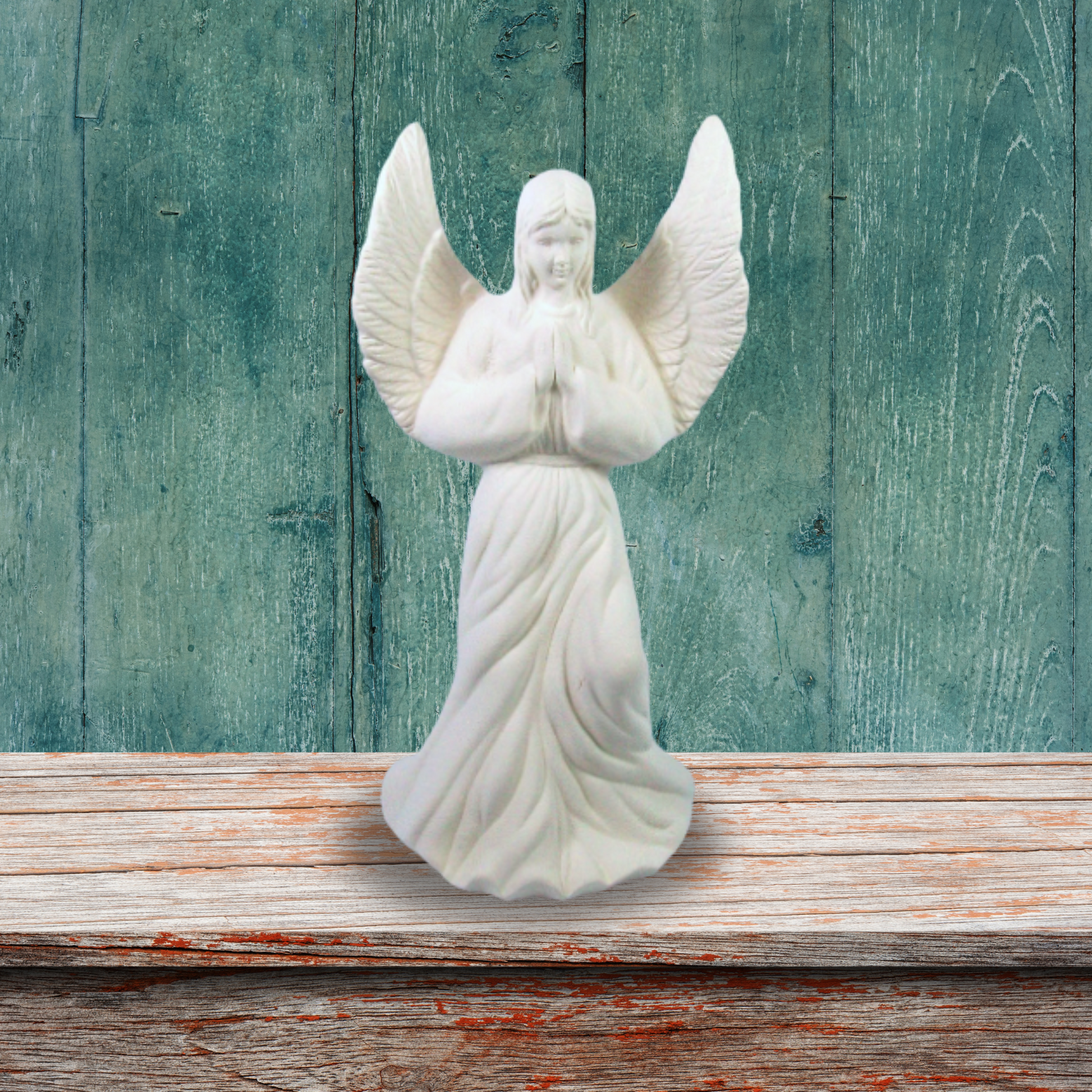 Unpainted angel standing on a brown shelf in front of a rustic green wooden wall.  Her wings are outstretched and her hands are in the prayer position