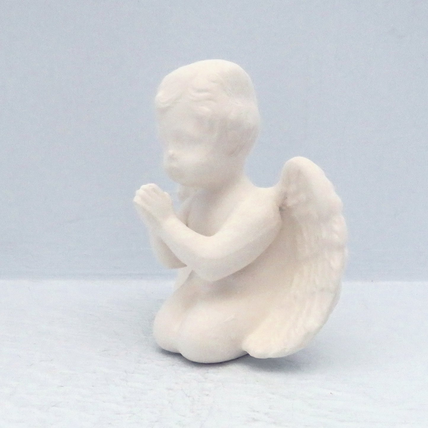 side view of paintable ceramic praying cherub kneeling on a blue wood shelf