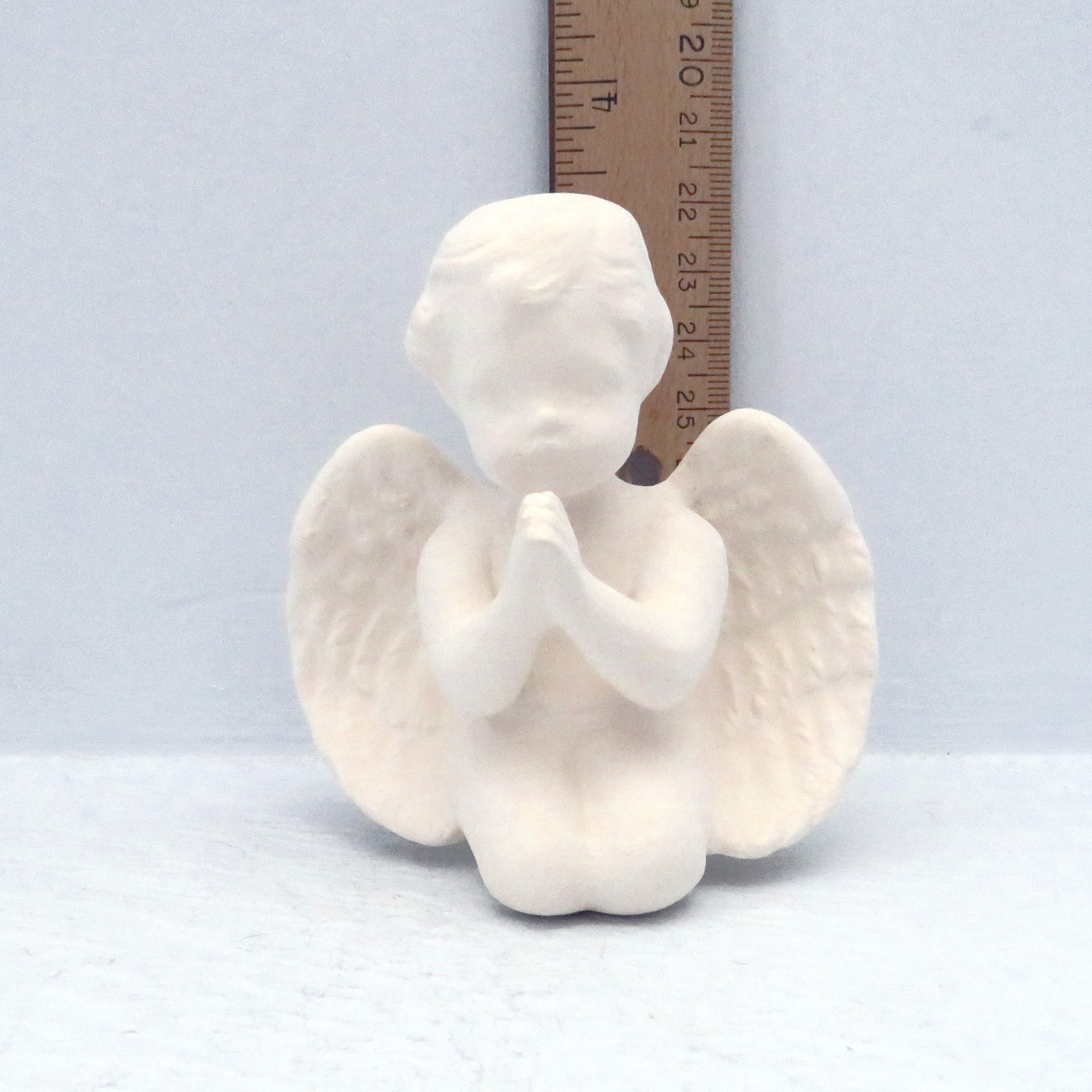 Ready to paint handmade ceramic cherub kneeling with hands folded in prayer position by a ruler showint it to be 3 1/4 inches tall