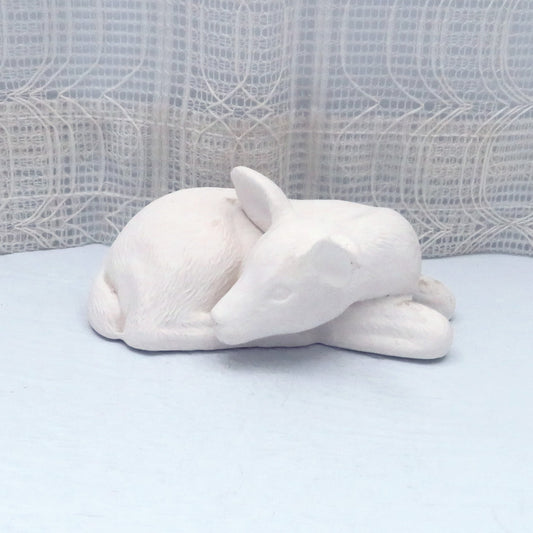 handmade ready to paint ceramic reindeer figurine lying on a blue surface with a lacy curtain behind it.  His legs are curled underneath him and his heasd is looking backward, with his head resting on his body