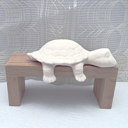 Ready to paint ceramic shelf turtle on a wooden pedestal with a lacy curtain behind.  The head is on the pedestal and the right legs are dangling off.