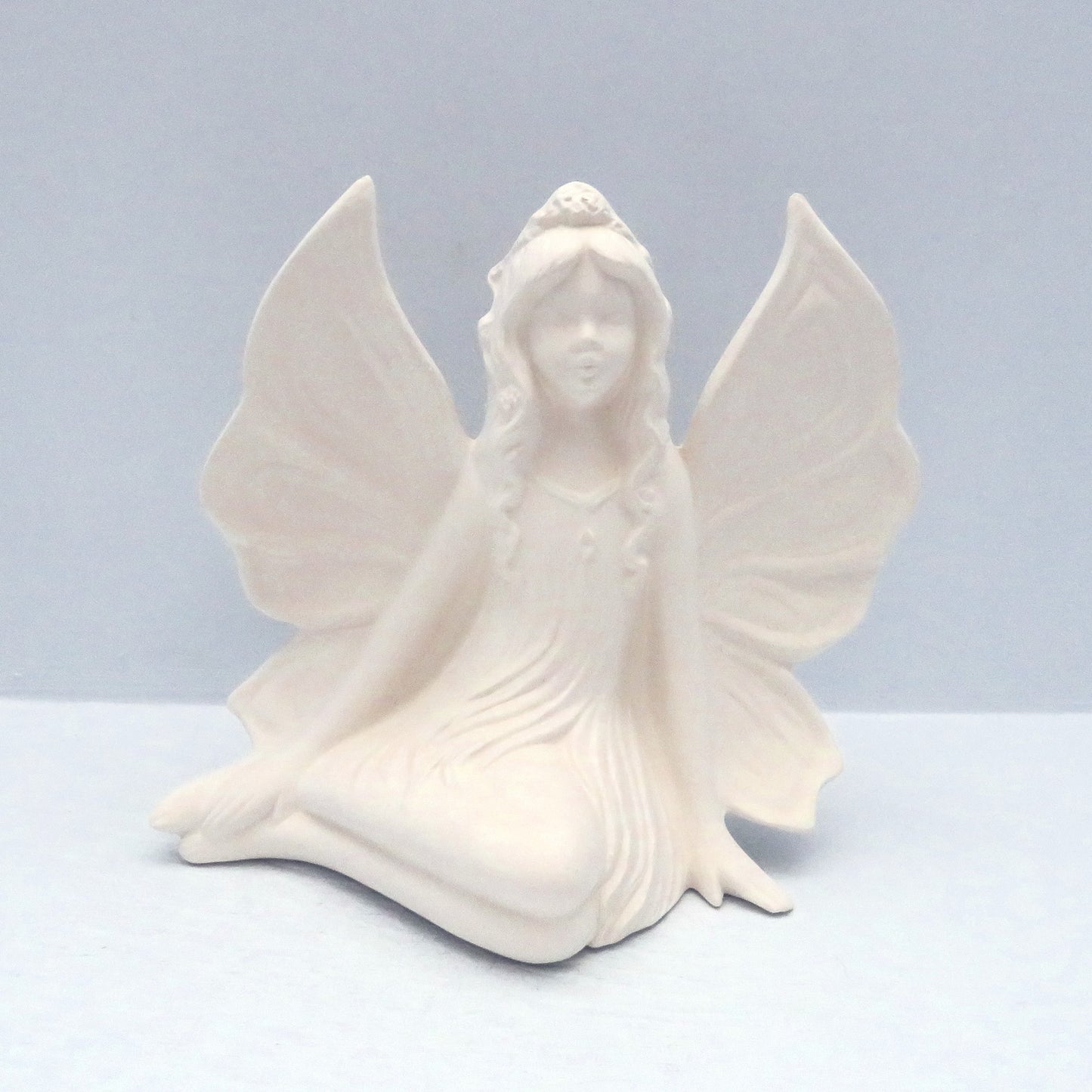 Handmade paintabla ceramic side sitting fairy sitting with her wings out facing the camera againsta a blue surface.