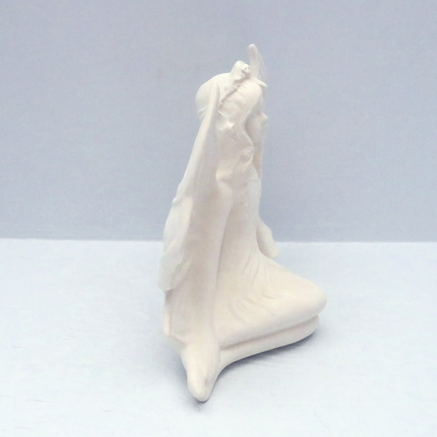 Handmade unpainted bisque fairy figurine facing to the right sitting with her legs to the right on a plae blue surface