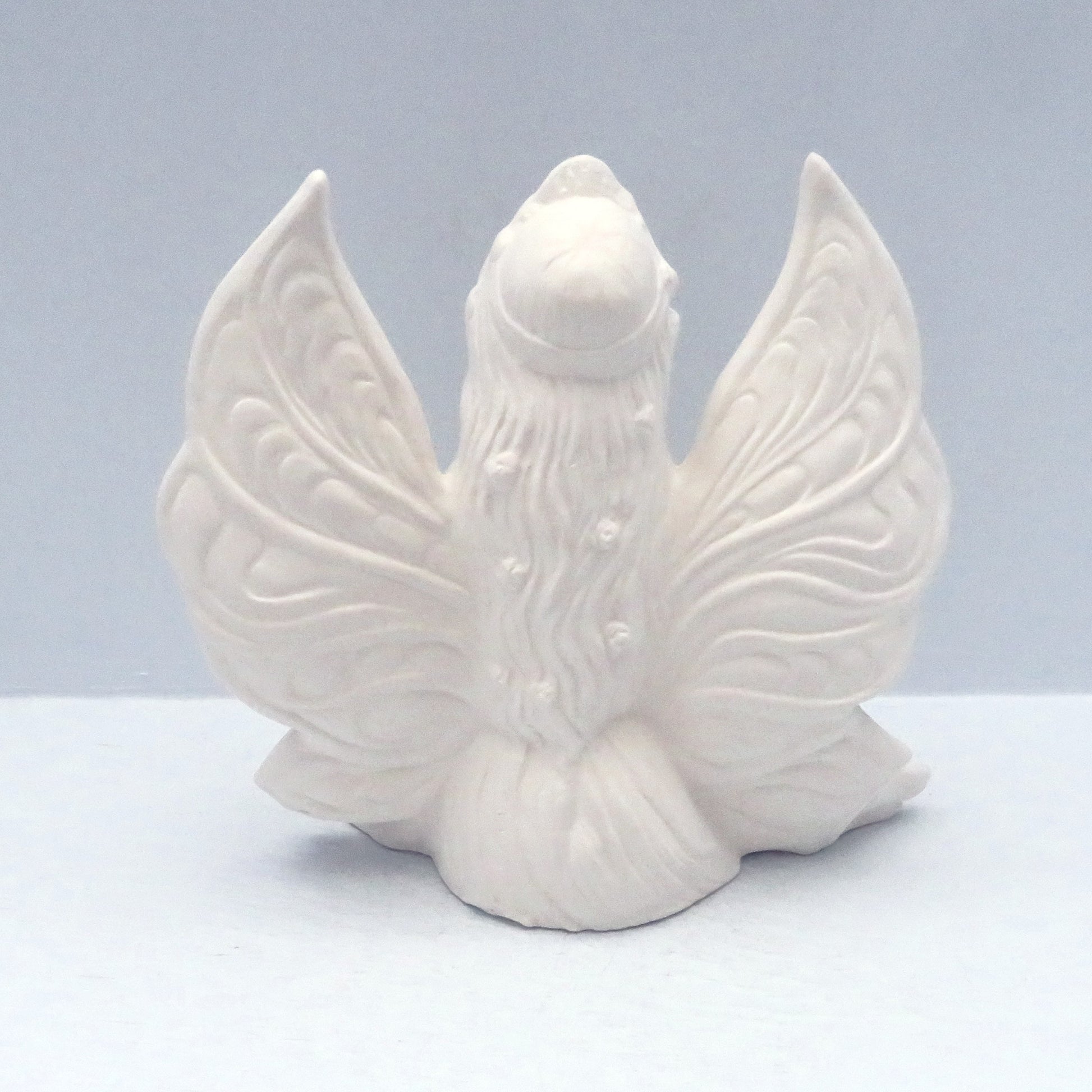 Rear view of unpainted ceramic fairy statue showing her long hair with flowers in it, the detail in her wings, on a light blue surface.