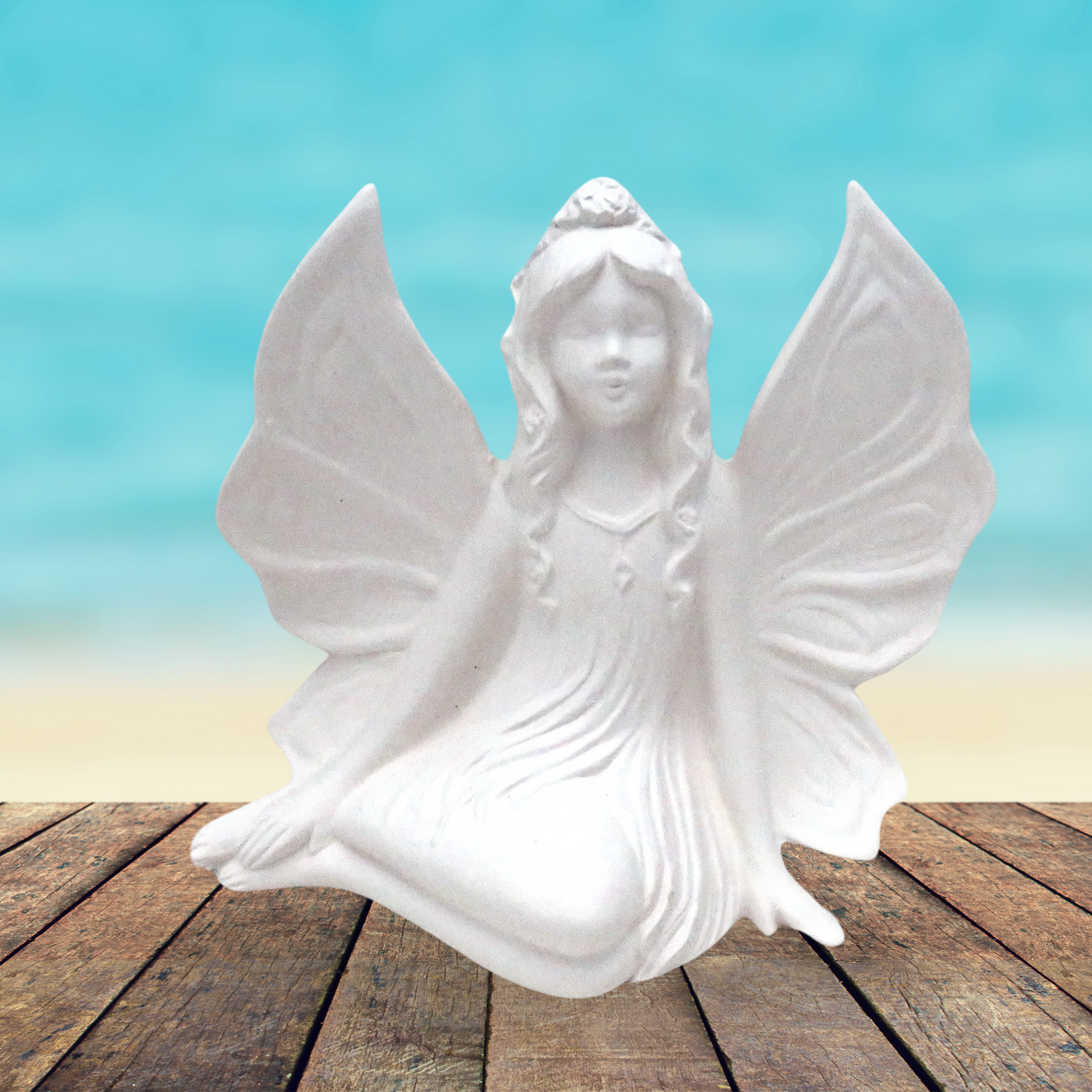 handcrafted ready to paint ceramic fairy sitting on a deck by the ocean.  Her wings are outstretched and her legs are curved to her right.  One hand is on the table and one is on her feet.