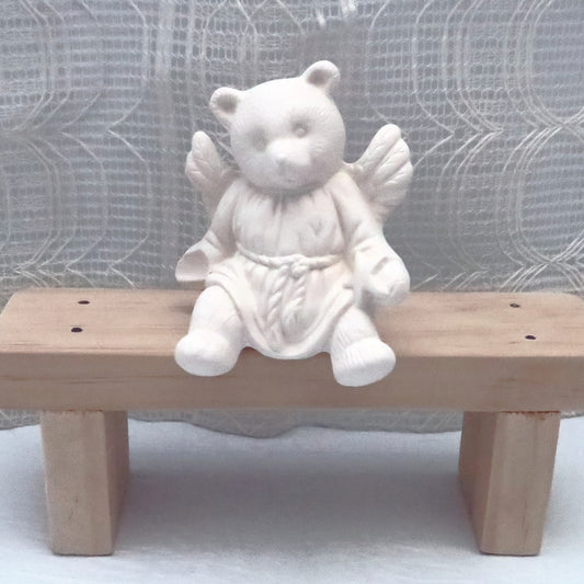 Ready to paint ceramic angel bear figurine sitting on a wooden bench.  Its wings are outstretched.  There is a star in one hand and a place to affix a stone or flower in the other hand.