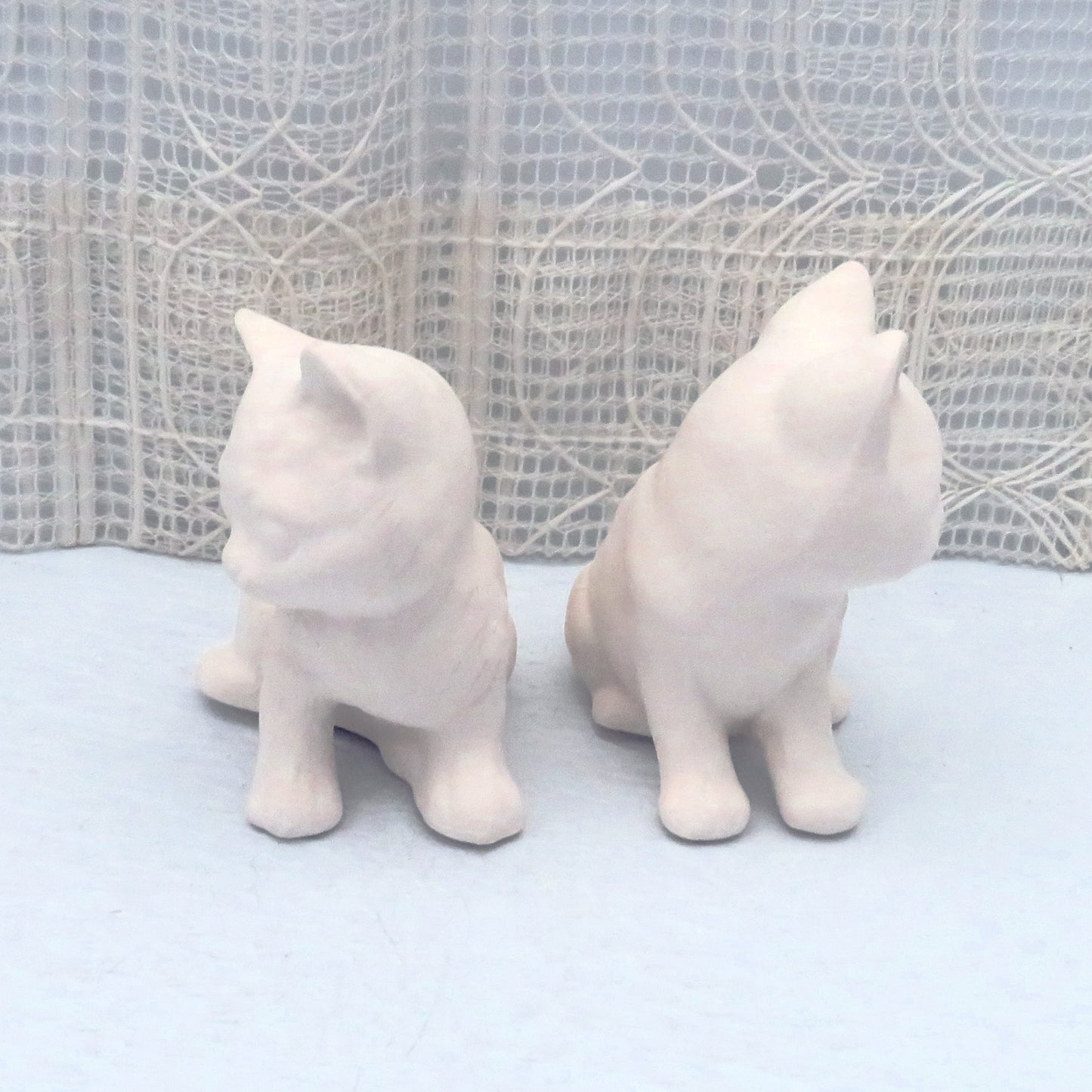 2 sitting ceramic cat figurines with the fronts forward and their faces turned away from each other.  They are on a blue table and have a lacy ecru  background.