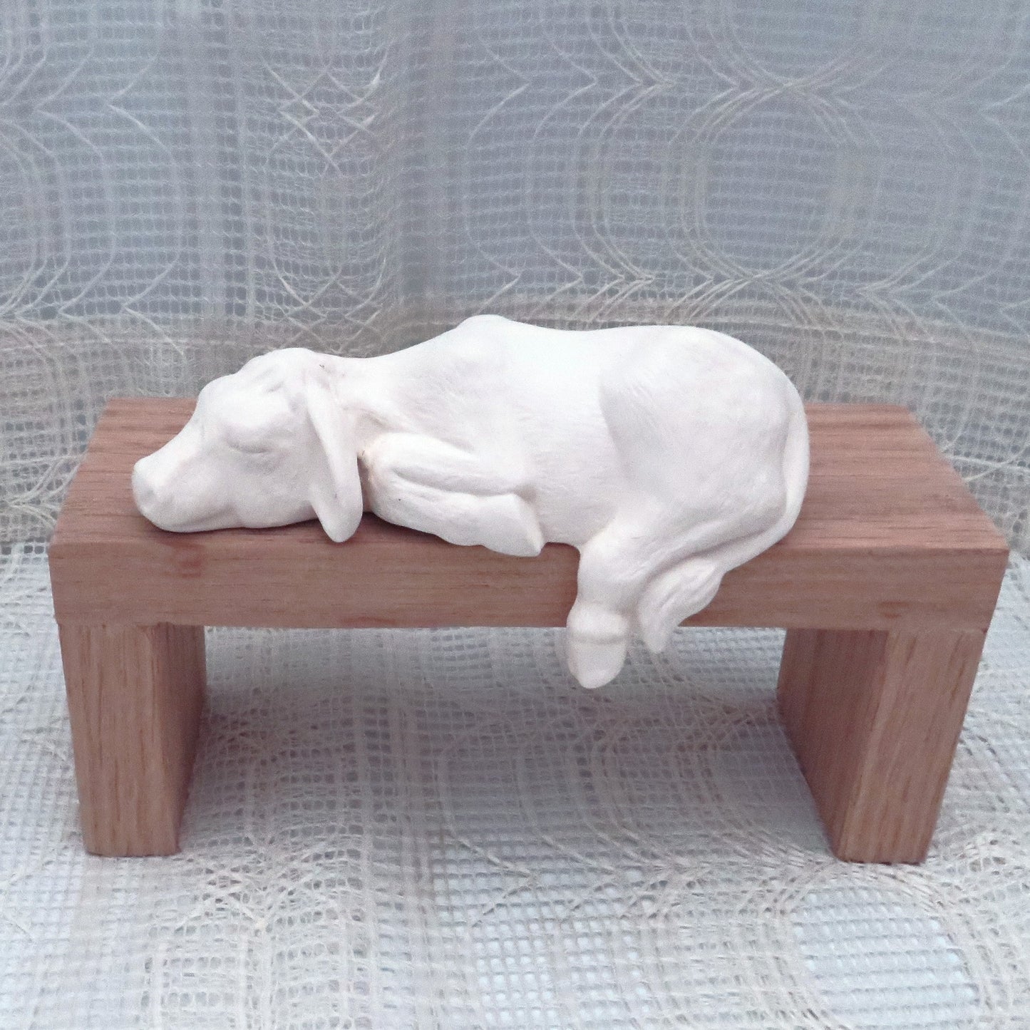 Paintable ceramic shelf cow sitting on a wooden platform with the lacy curtain and cloth behind and under.