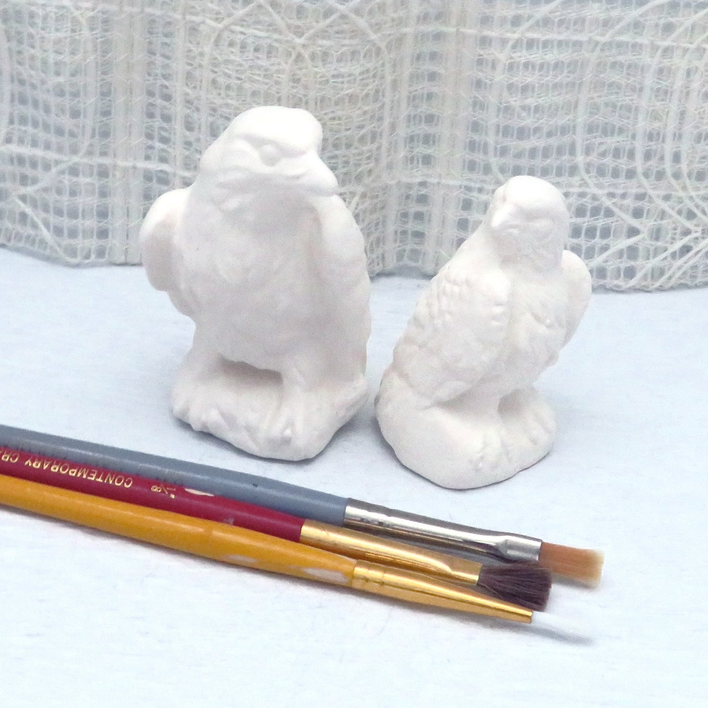 2 small unpainted ceramic eagle figurines on a pale blue table with an ecru lacy curtain.  There are 3 paintbrushes in front of the eagle figurines.  This is a front view of the eagles.