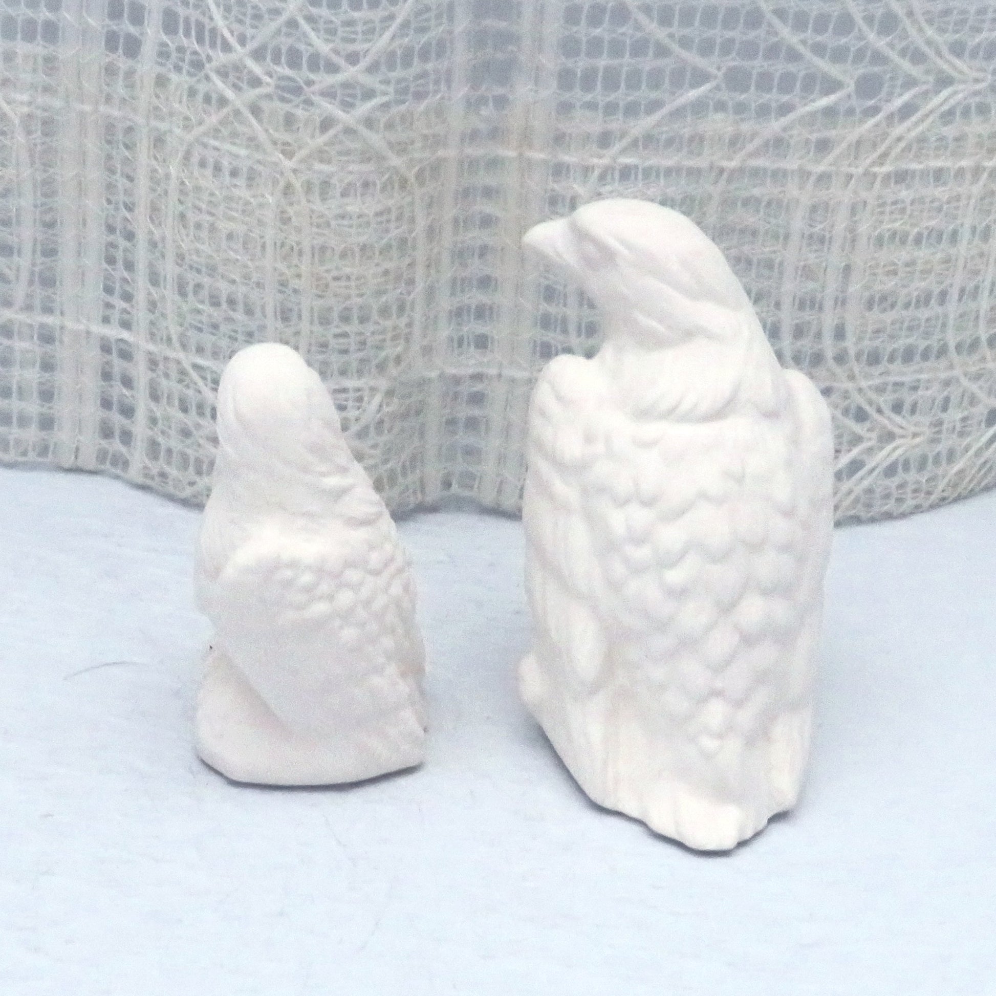Rear view of ready to paint ceramic eagle figurines ona pale blue table and an ecru lacy curtain.