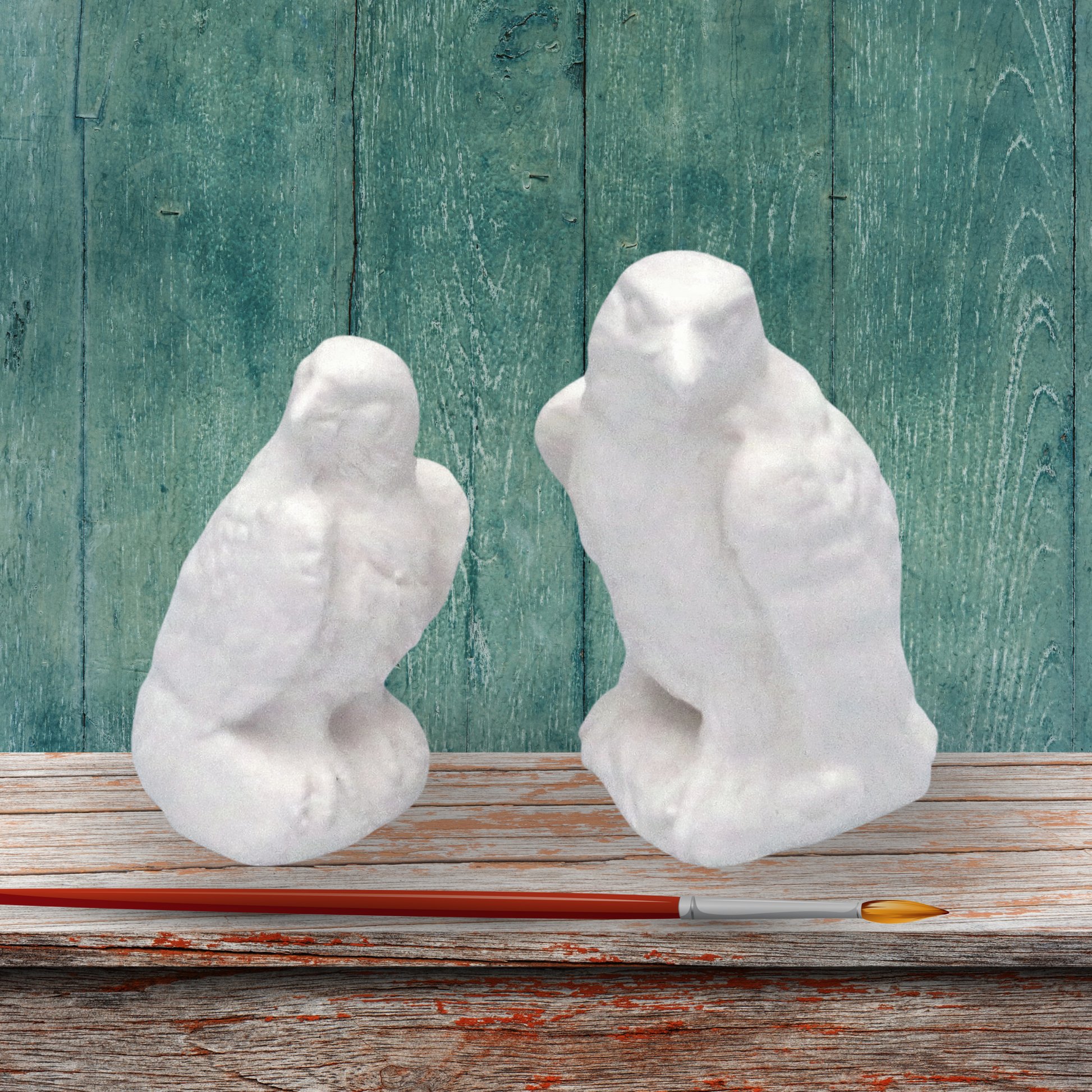 2 small unpainted eagle statues on a rustic wood table with green background and a paintbrush in front of them.