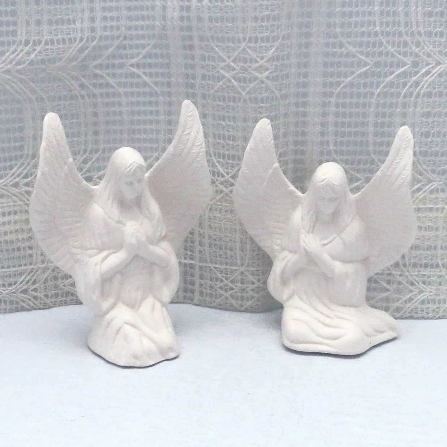 Handmade ready to paint ceramic angek statues on a blue tablle with an off white lacy curtain behind.  The angels are facing forward