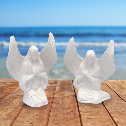 Set of 2 unpainted ceramic angel figurines sitting on a wood table by the ocean.  Both of the angels have their wings oustretched.  One is sitting side sitting, one is kneeling.