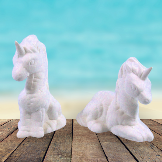 Set of 2 handmade ready to paint ceramic 'softy' unicorns sitting on a brown wooden table by the ocean.  One is sitting up, the other lying down.