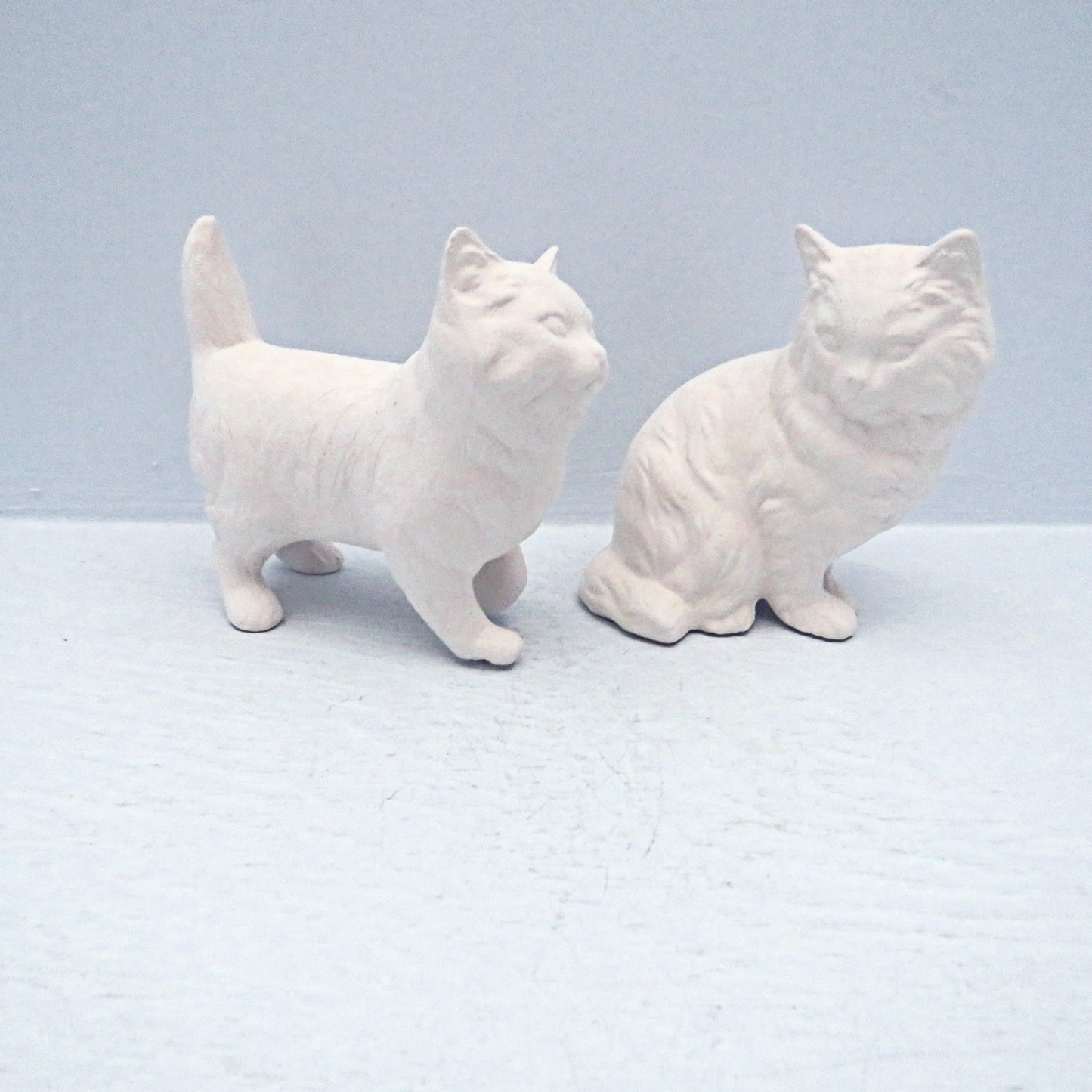 2 ready to paint ceramic cat figurines.  The one on the left is standng and  facing  the right.  The one on the left is sitting with its  body toward the right, and its head facing the camera  The standing cat has its tail pointing up.  They are on a pale blue surface.