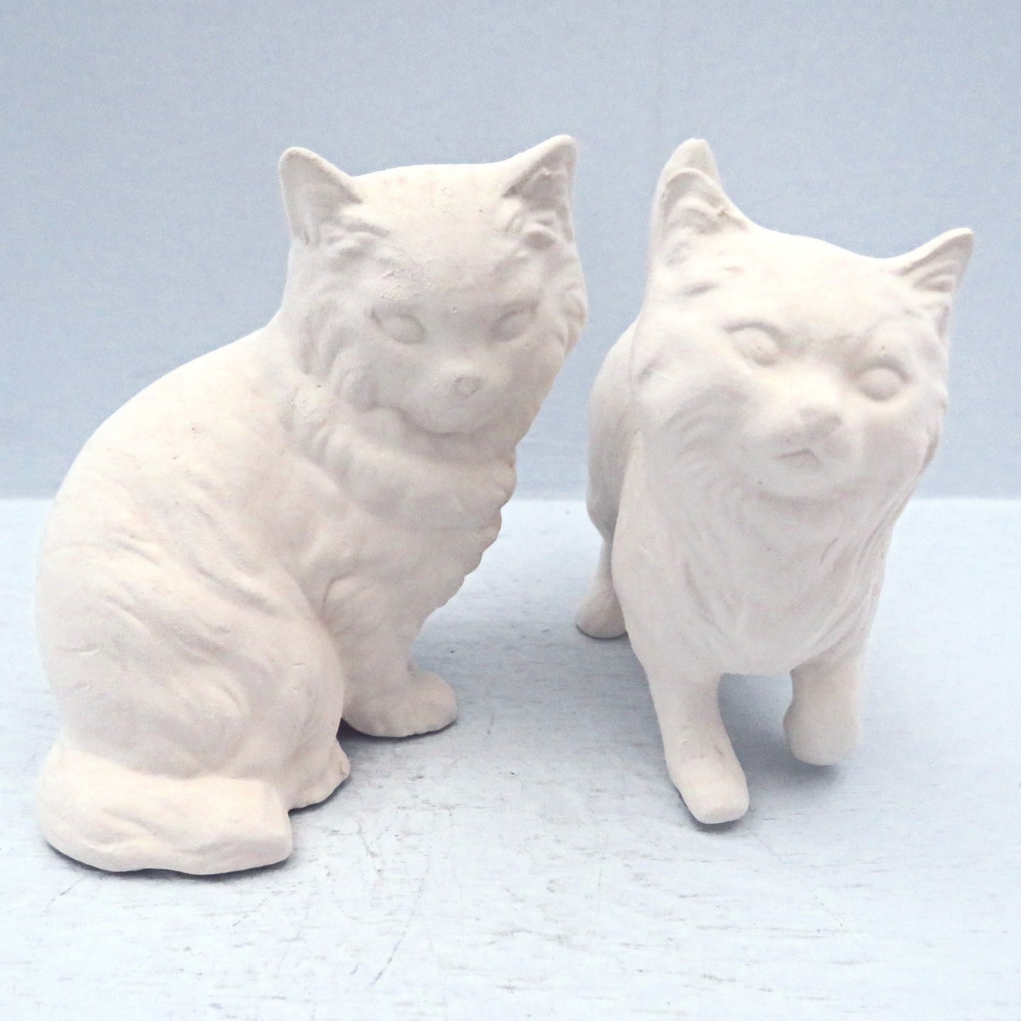 Set of 2 Unpainted ceramic cat figurines.  One is sitting, the other standing on a pale blue surface.  