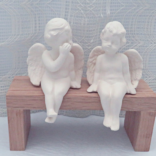 Set of 2 handmade paintable ceramic cherub figurines sitting on a bench in front of an ecru lacy curtain.  The cherub on the left has his hand covering his mouth and is facing the one on the right.  The ready to paint cherub statue on the right has his head cocked as if he is listenting to the other.