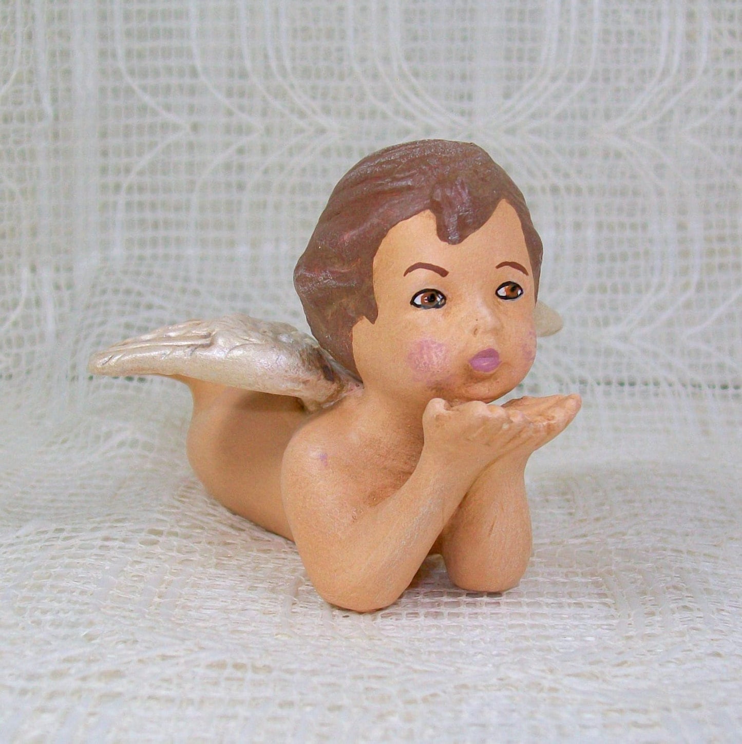 Handmade Ceramic Cherub figurine on tummy blowing kisses on a lacy table in front of a lacy curtain.