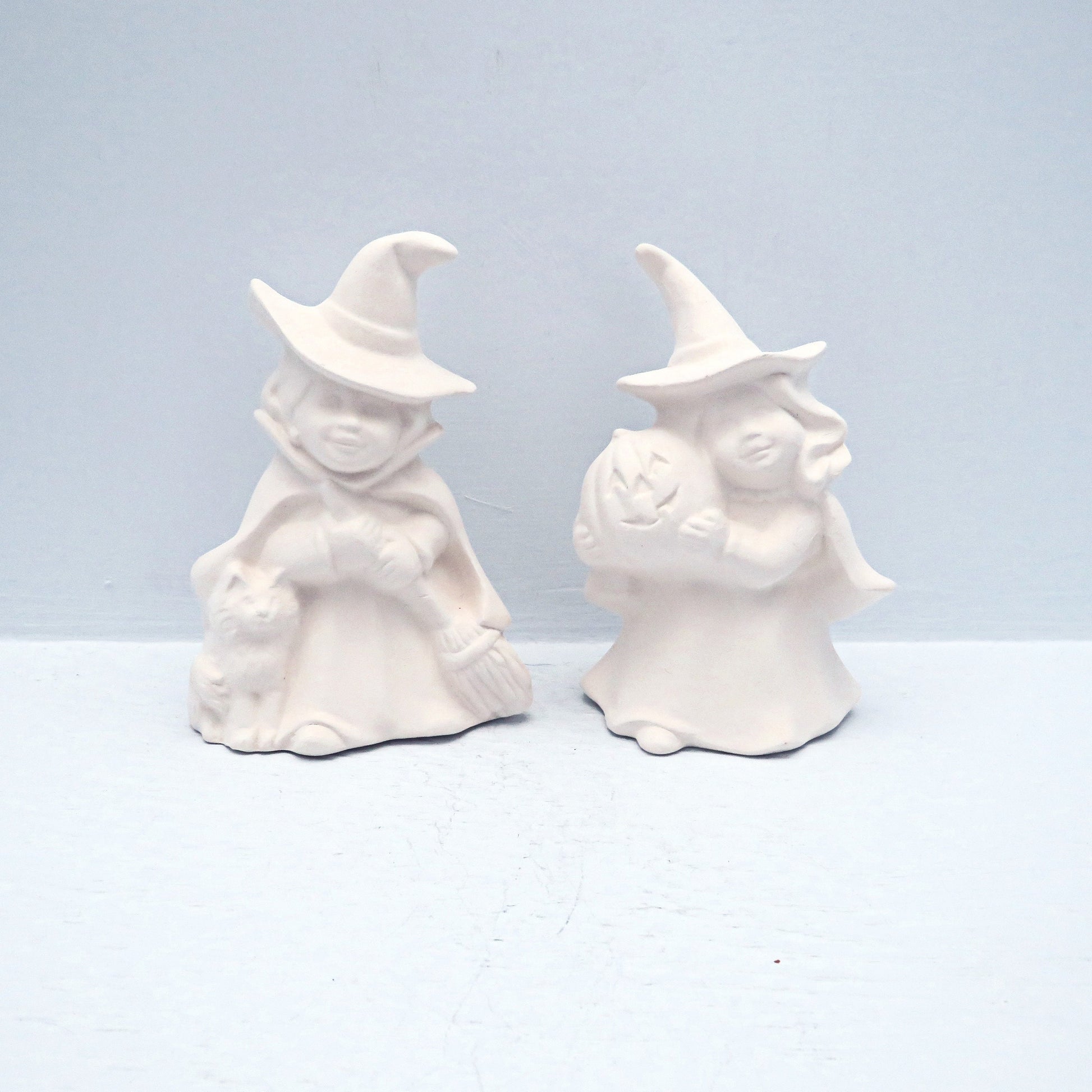 Set of 2 ready to paint ceramic witch figurines standing on a pale blue surface.  One is holding a pumpkin, one has a cat and a broom.