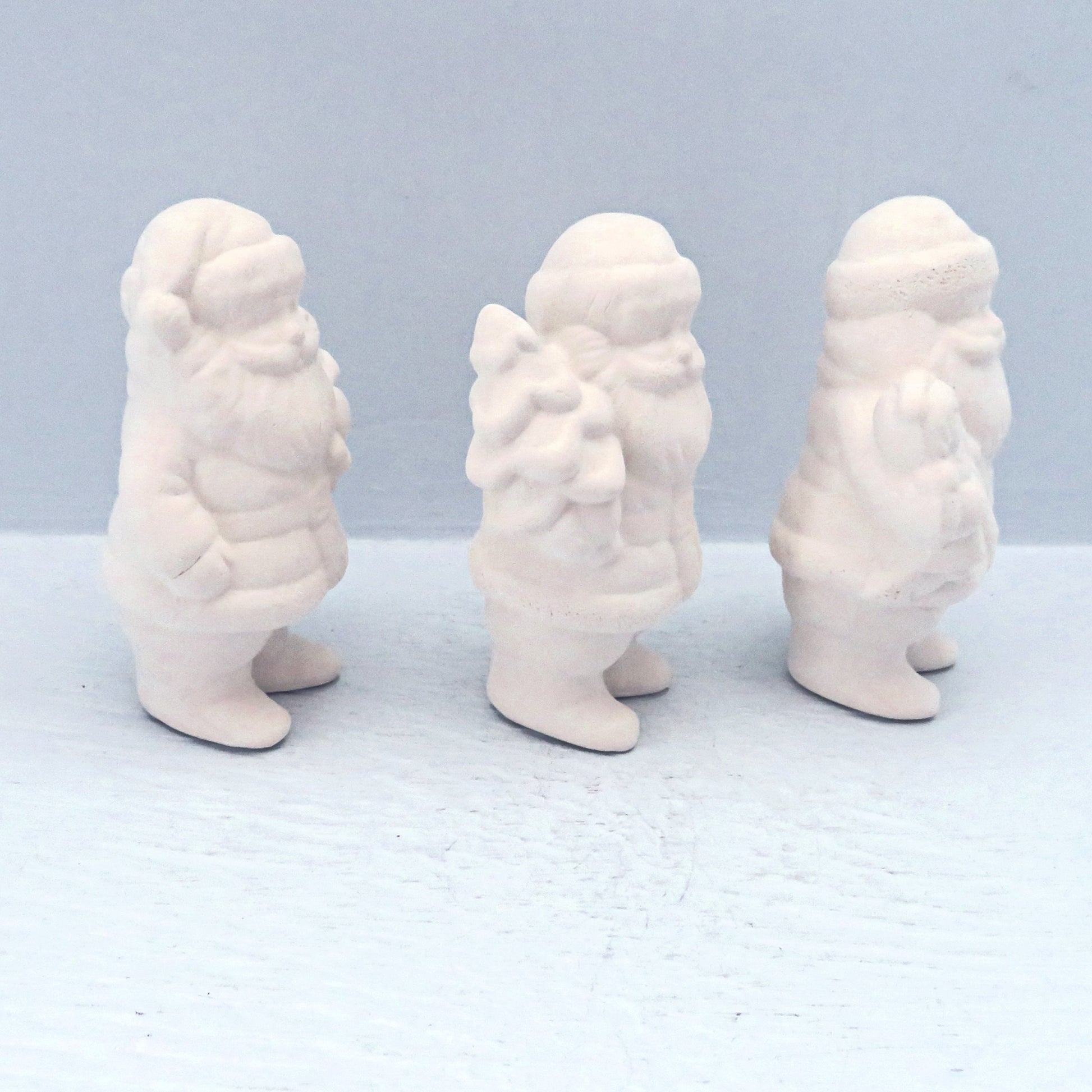 Side view with paintable ceramic Santa statues facing right on a pale blue surface.