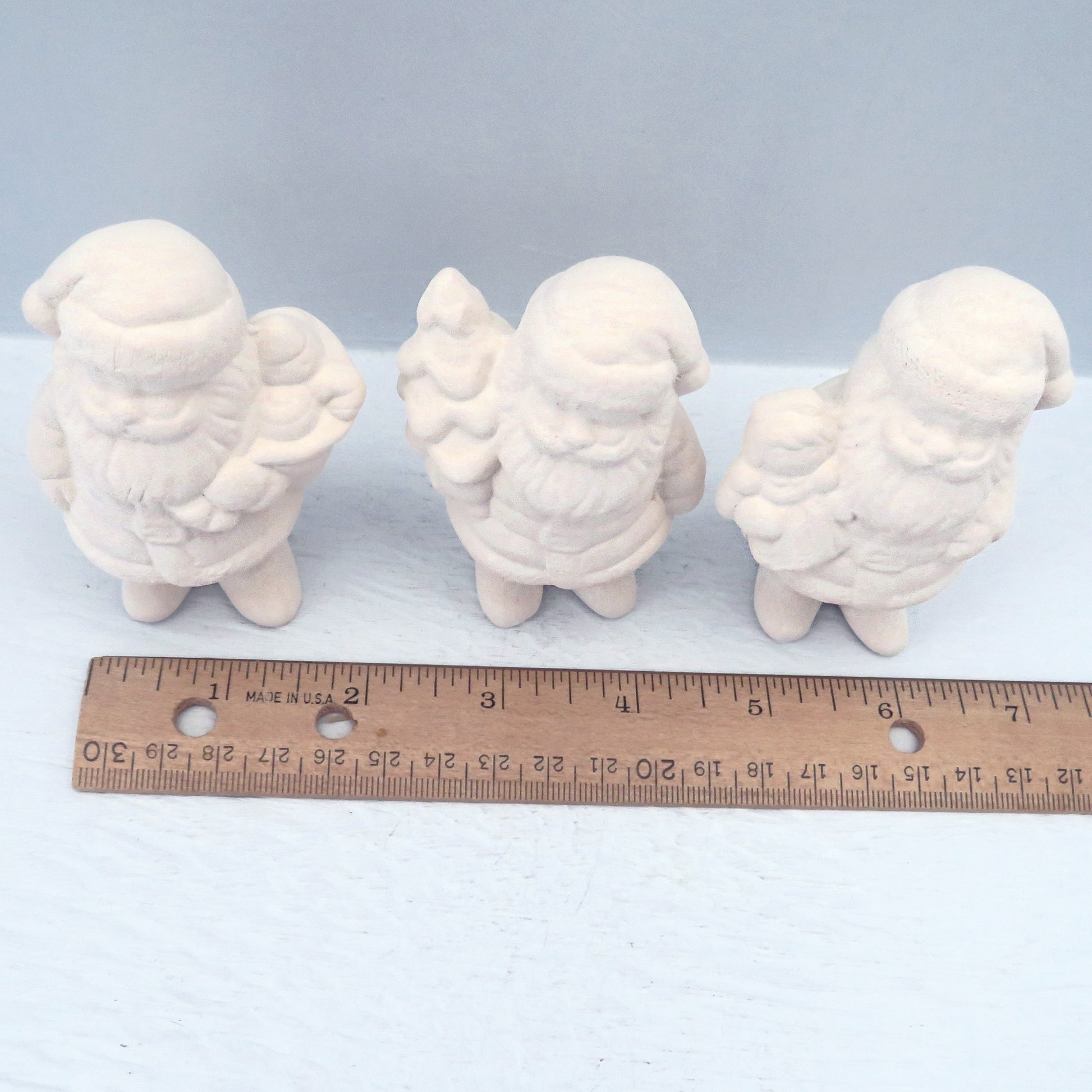 Unpainted bisque santa claus figurines standing near a ruler showing them to be approximately 1 1/2 to 1 3/4 inches wide