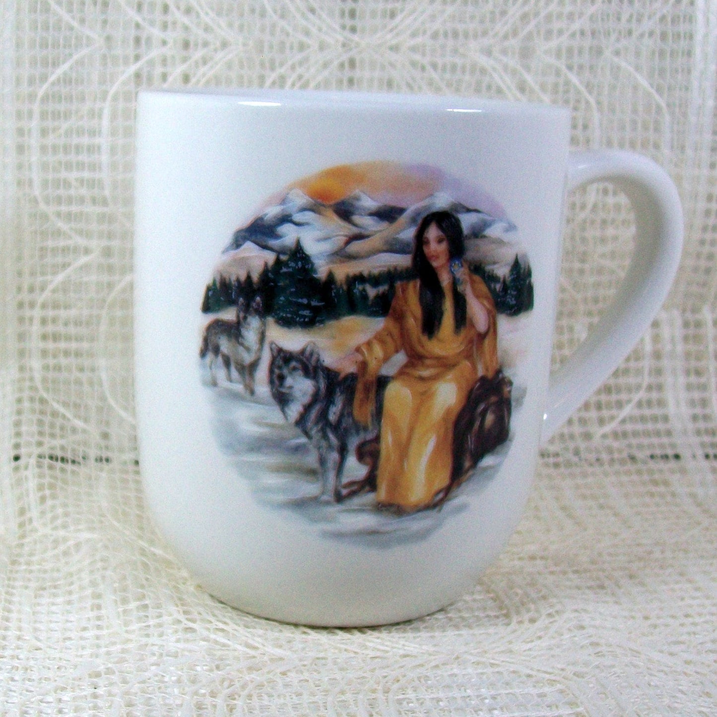 white glossy ceramic mug on a lacy background.  The handle s facing right.  This view shows a close up of the Native American girl sitting with a Southwest background including 2 wolves.