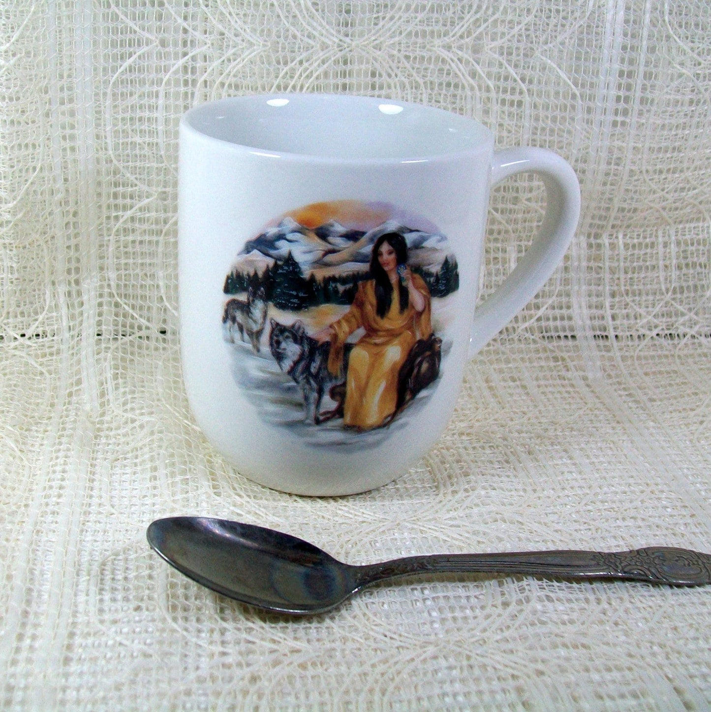 White glossy mug with native American girl.  The mug is on an off wihite lacy cloth and there is a tea spoon in front of it for size comparison