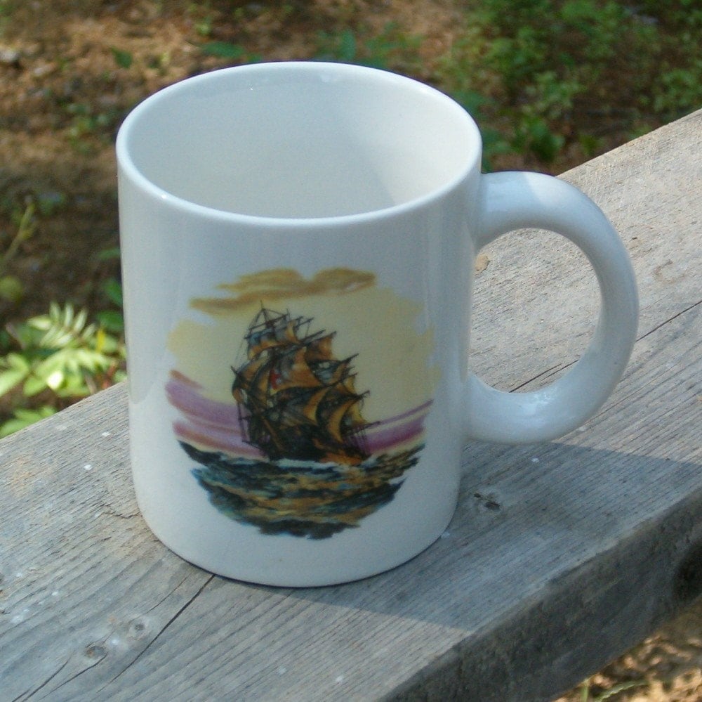 Front side of white ceramic mug with a clippe ship and ocean scene against a sunset sky.  The mug is sitting on a cedar shelf.