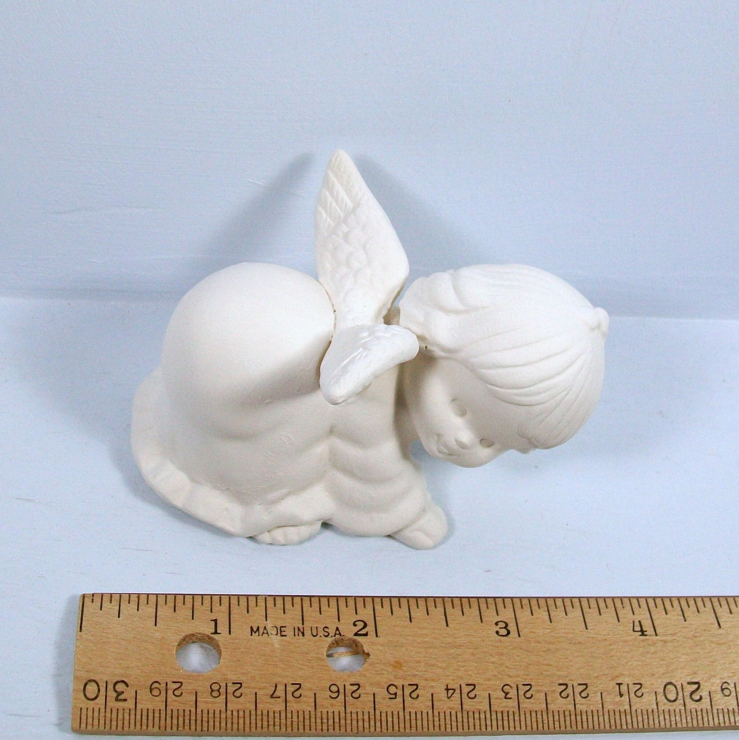 Unpainted ceramic angel statue on all 4's looking from above.  She has her wings outstretched and her head turned toward the right.  She is near a ruler showing her to be approximately 3 1/2 inches long.