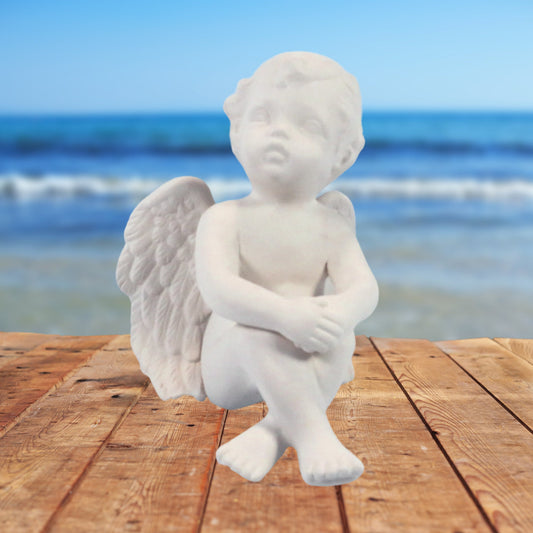 Ready to paint cherub figurine sitting on a talbe by the ocean.  Its knees are up, feet crossed, and his hands are clasped and resting on his knees.