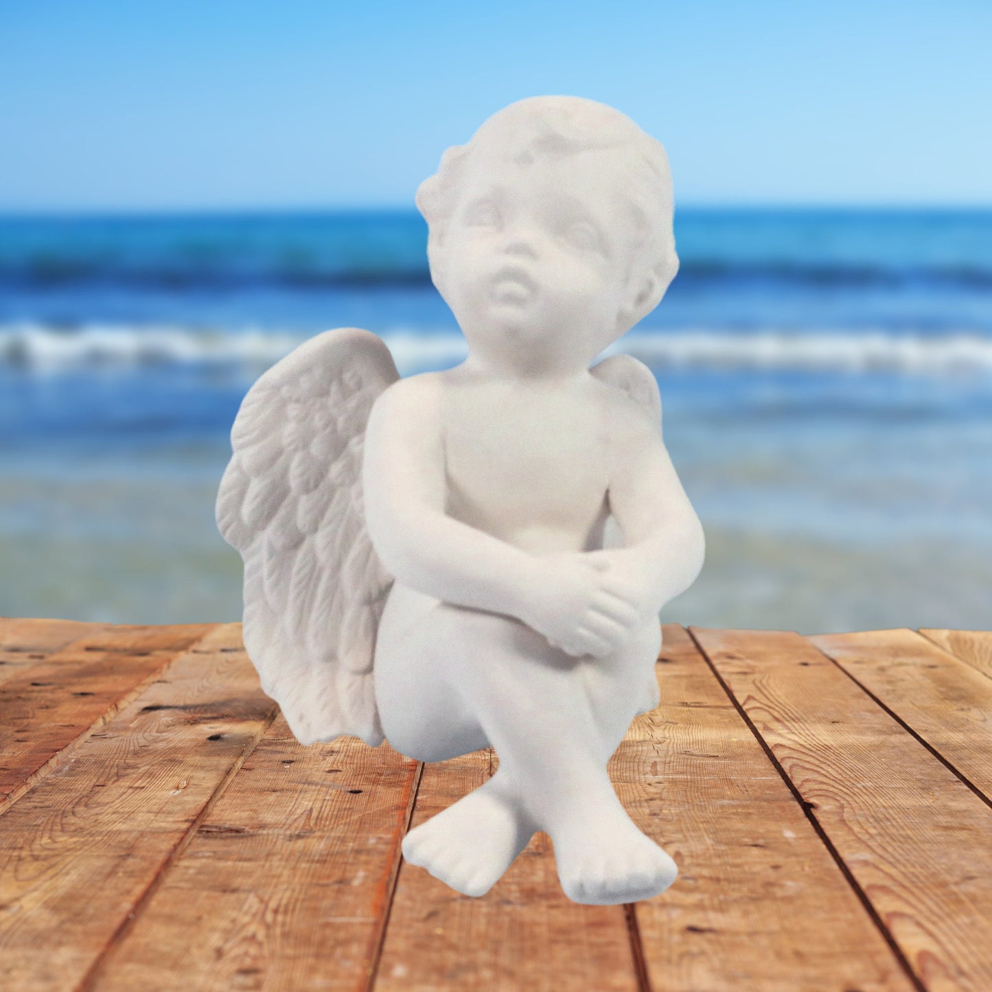 Ready to paint cherub figurine sitting on a talbe by the ocean.  Its knees are up, feet crossed, and his hands are clasped and resting on his knees.