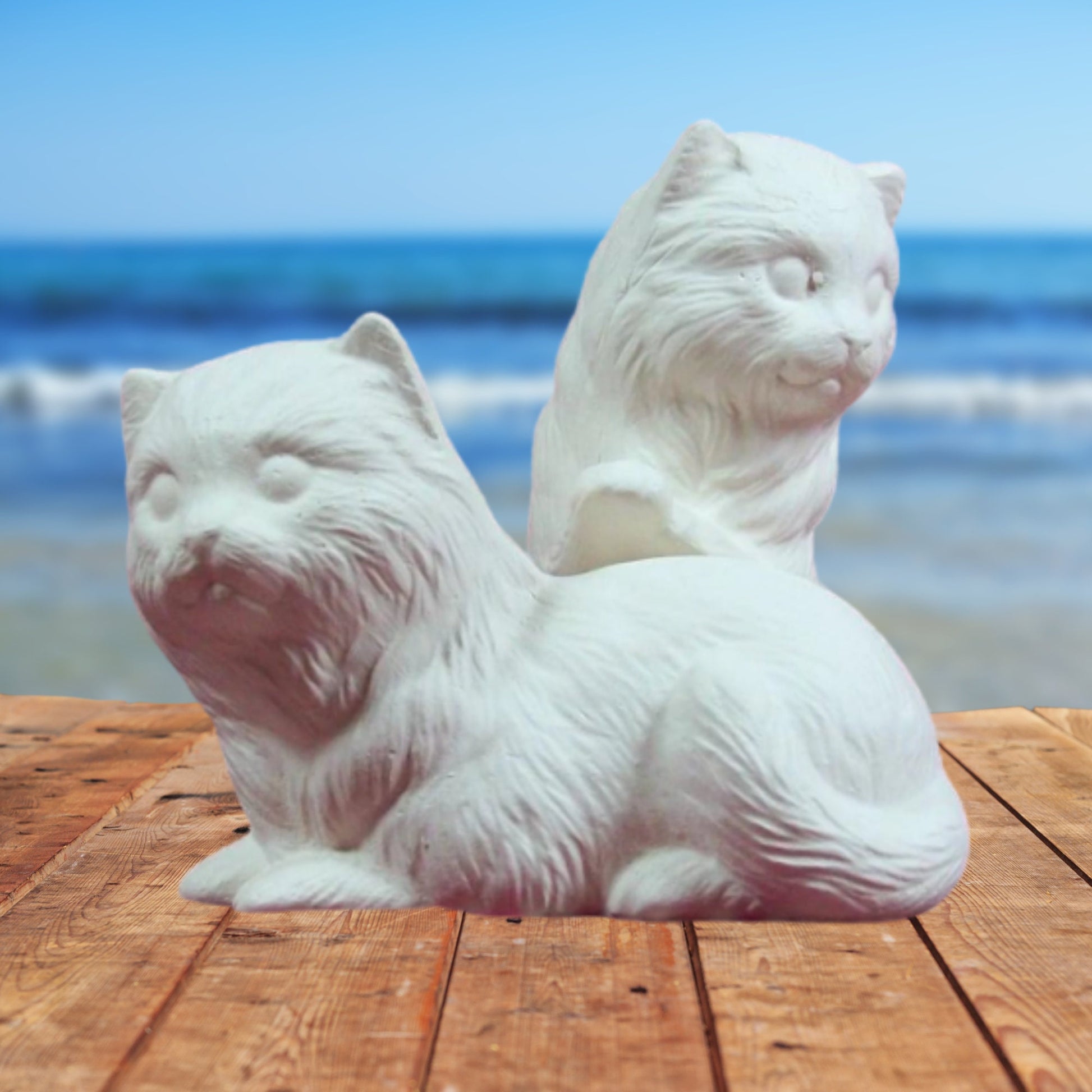 2 ready to paint ceramic figurines on a table by the ocean.  One of the cats is lying down, The other is jumping with its front feet on the back of the first one.  