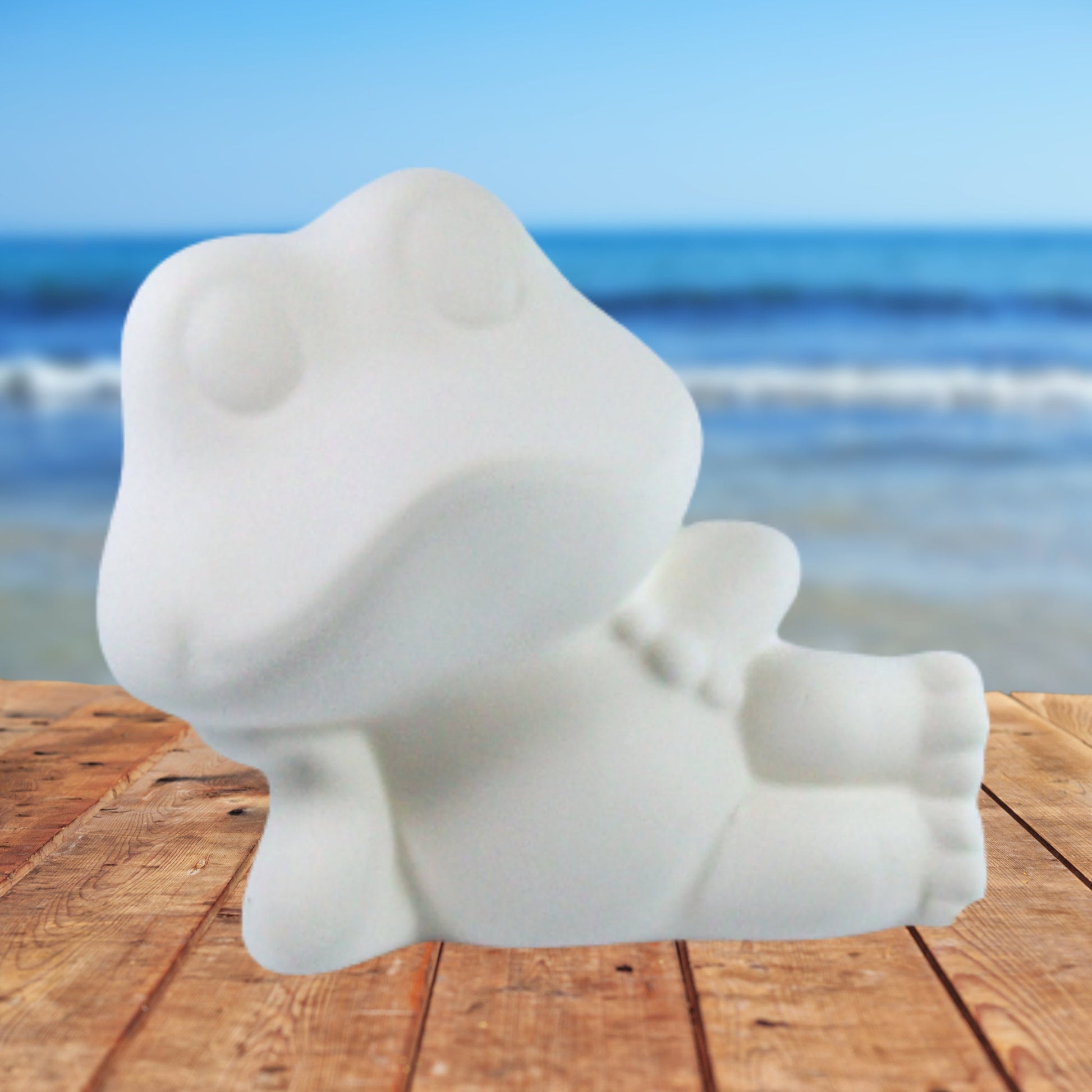 Handmade ready to paint ceramic frog lying on his right side with his head resting on his right hand lying on a wood table by the ocean.