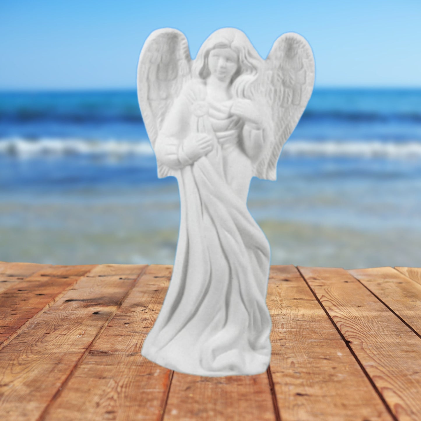Standing ready to paint ceramic angel with long robes with her wings tucked in.  Her left hand is to her chest, the right hand is on her trunk.  She si standing on a table by the ocean.